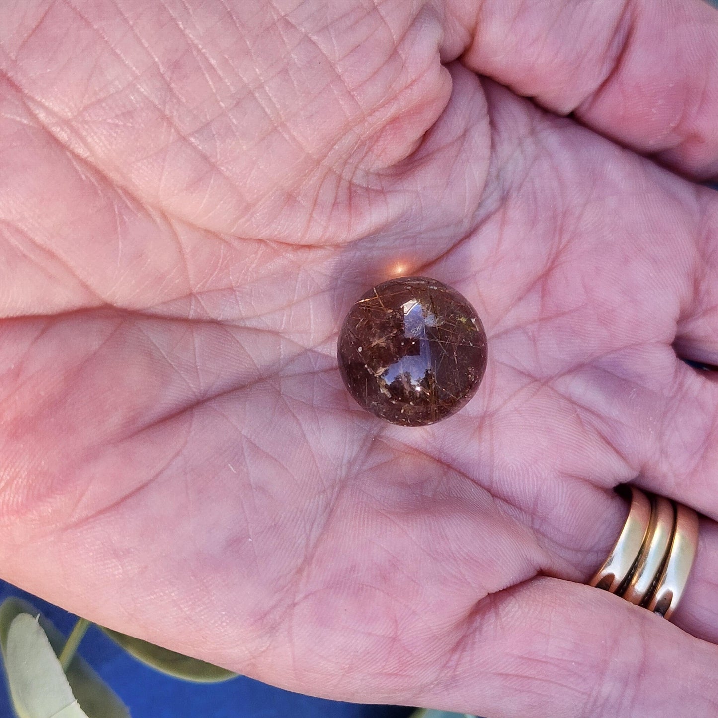 A close-up image of an open palm holding The Crystalary's Rutile Quartz mini sphere 15mm. The person's hand has prominent lines, and they are wearing a gold ring on their ring finger. The background shows a hint of greenery, setting the perfect scene for meditation and reflection.