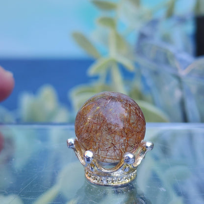 A Rutile Quartz mini sphere (15mm) from The Crystalary, featuring clear quartz with golden rutile inclusions, is elegantly positioned on a silver crown-shaped stand. The backdrop is softly blurred with shades of green and blue, accented by hints of foliage, creating an enchanting and sophisticated ambiance.