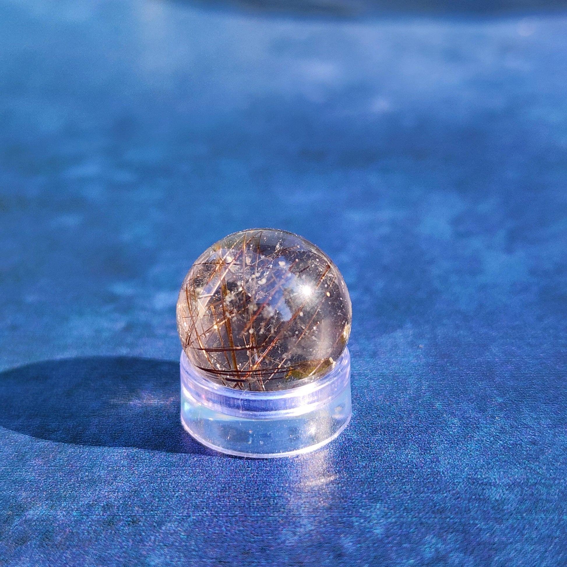 A Rutile Quartz mini sphere 15mm from The Crystalary, featuring fine, thread-like inclusions, is placed on a clear plastic stand. The background is a smooth, vibrant blue surface that enhances the clarity and detail of the polished crystal, making it perfect for meditation.