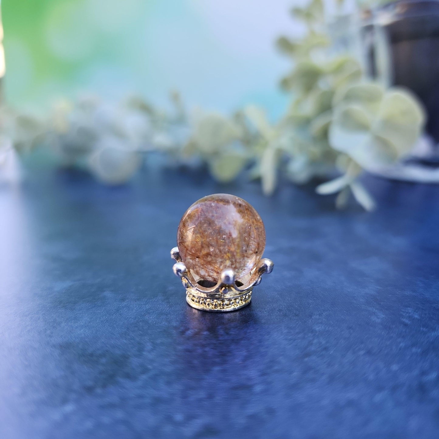 A small, spherical Rutile Quartz mini sphere 15mm from The Crystalary is placed in a decorative stand resembling a golden crown and showcased on a smooth, dark surface. The background is softly blurred, featuring light green foliage, giving the image a tranquil and elegant aesthetic.