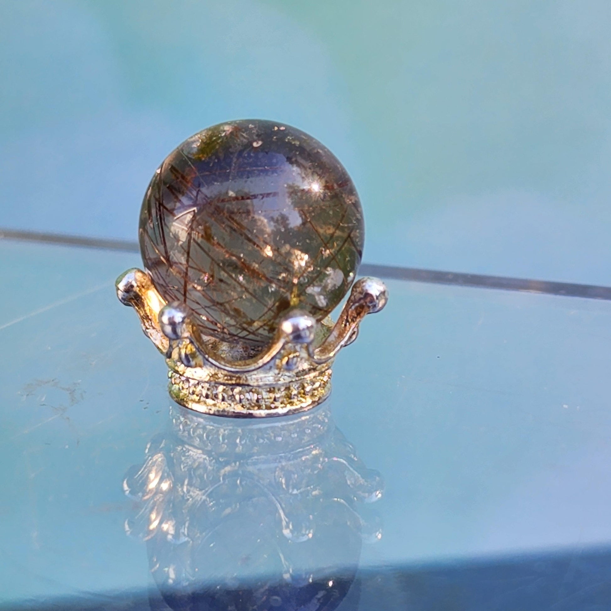 A Rutile Quartz mini sphere 15mm from The Crystalary, polished to perfection and featuring golden inclusions, sits elegantly in an intricate, ornate gold stand shaped like a crown. The setup is set against a blurred blue background, with the surface below reflecting both the sphere and the crown for a stunning mirrored effect.