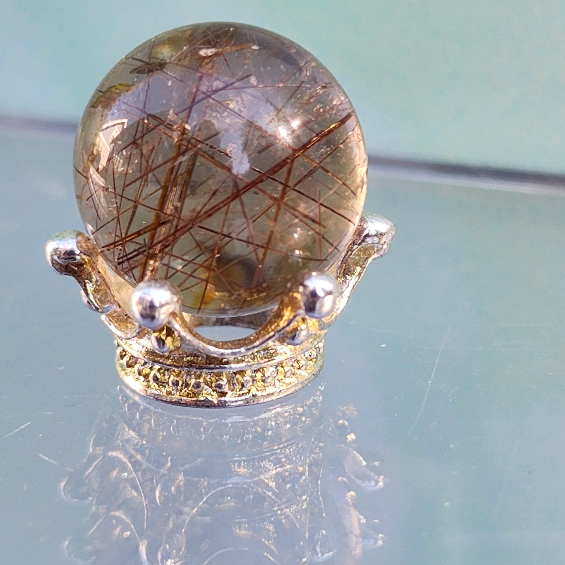 A polished Rutile Quartz mini sphere 15mm from The Crystalary, showcasing needle-like inclusions, rests on an ornate silver crown-shaped stand adorned with small yellow gemstones. Perfect for meditation, the backdrop is a soothing gradient of blue and green.