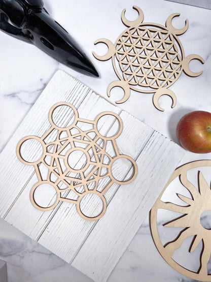 A flat lay photo showcases three intricate wooden cutouts from The Crystalary's Sacred Assorted Crystal Grids in Birch, featuring geometric and circular patterns reminiscent of sacred geometry designs. An apple is positioned near one of the cutouts on a white, marbled surface for contrast, evoking the feel of a crystal grid awaiting its crystal combinations.