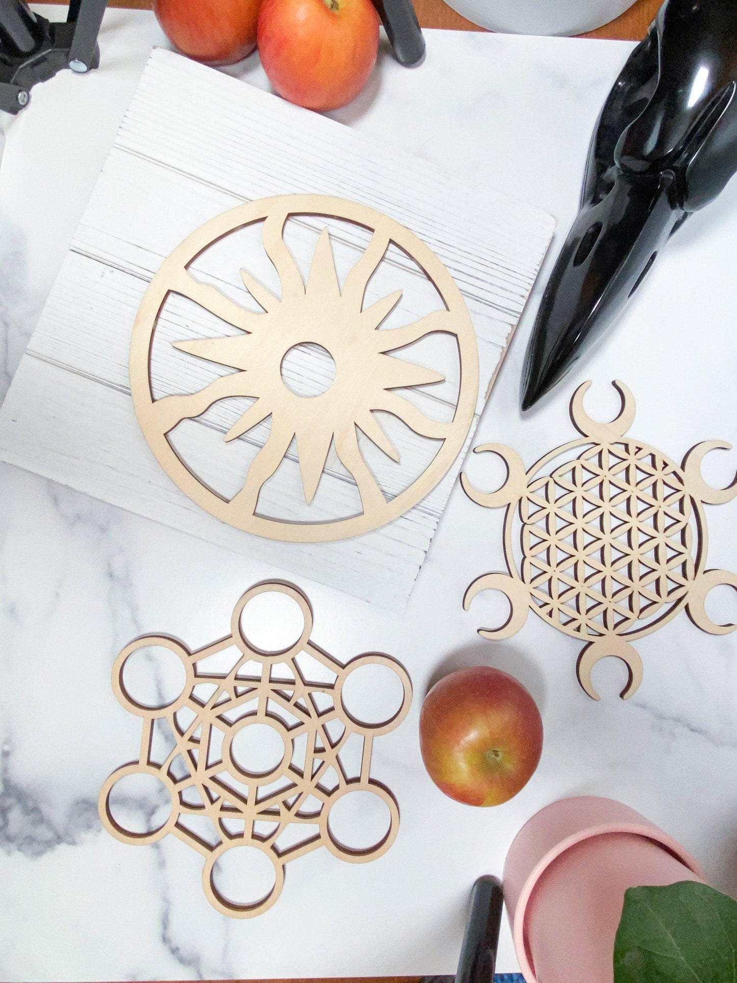 A top-down view of the marble surface showcases The Crystalary's Sacred Assorted Crystal Grids in Birch, featuring wooden geometric cutouts including a sun design, the flower of life pattern, and circular patterns. This sacred geometry arrangement is beautifully enhanced by crystal grid elements, two red apples, a black decorative object, and other harmonious accents.