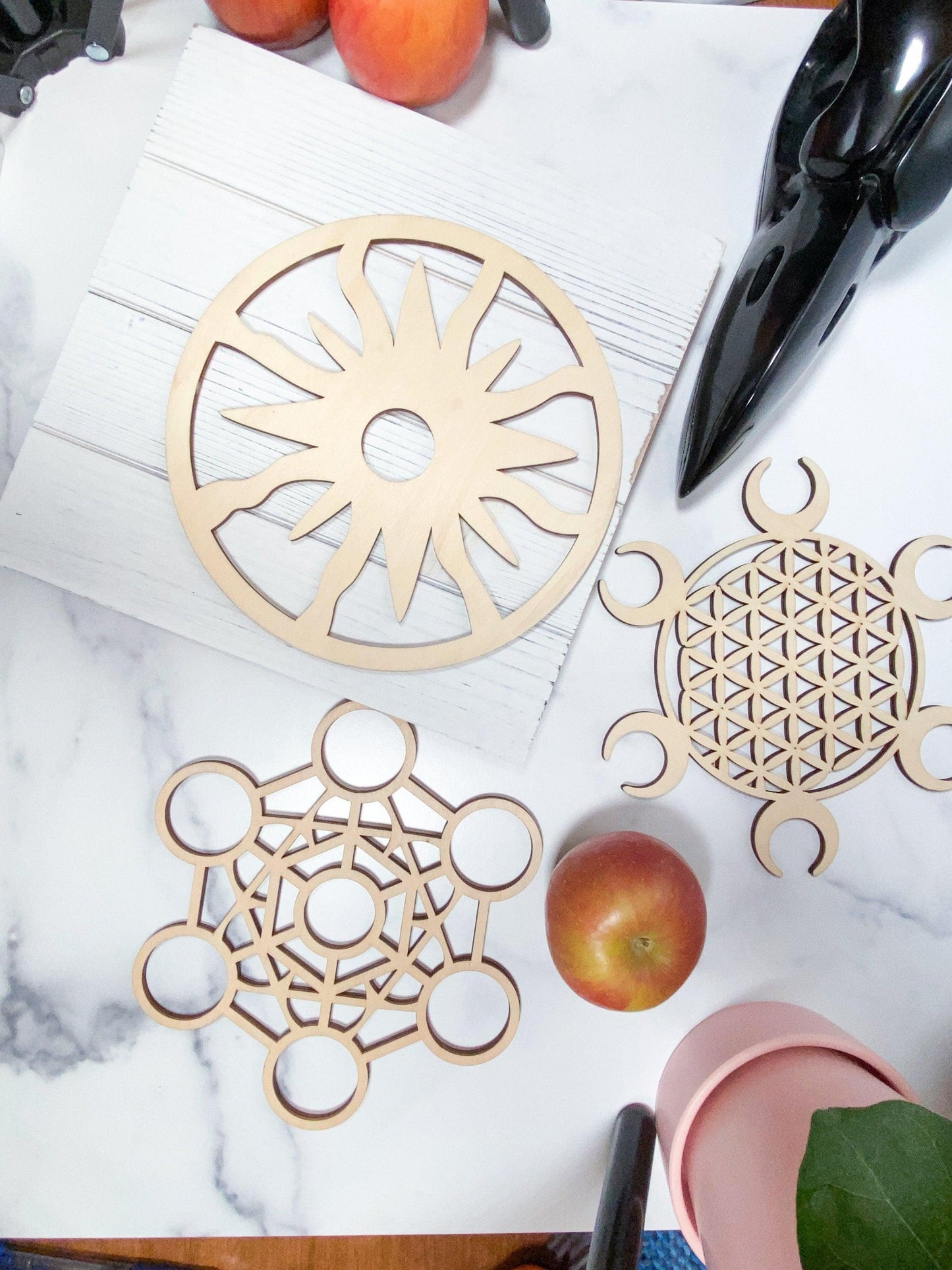 The image features three wooden geometric designs from The Crystalary's Sacred Assorted Crystal Grids in Birch collection on a white surface: a sunburst design in a circular frame, an intricate sacred geometry pattern with circles and lines, and a floral-like pattern with crescent shapes. Nearby, there is an apple, peach, and a black ceramic bird sculpture.