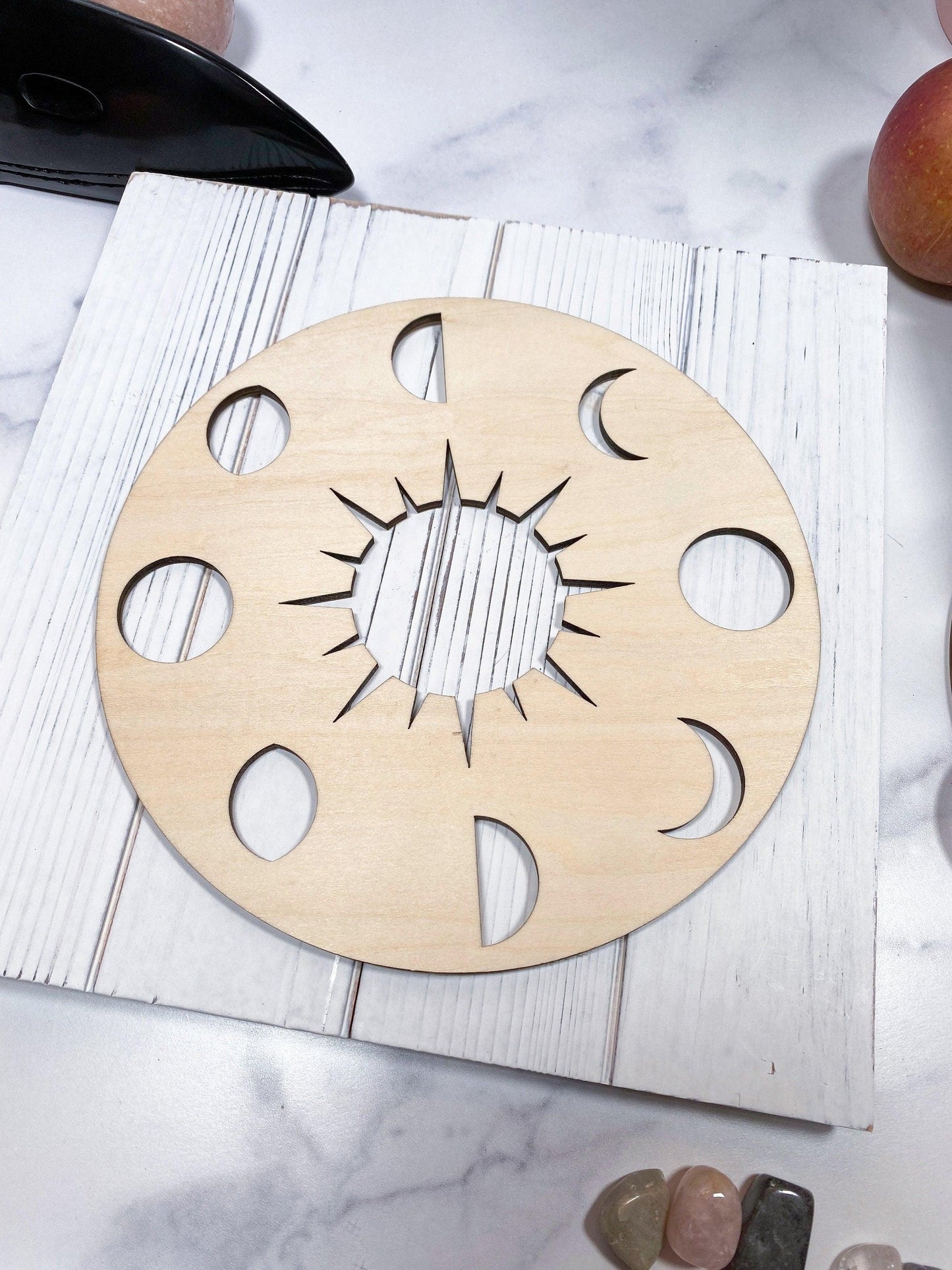 A circular birch cutout from 'The Crystalary,' showcasing the 'Sacred Assorted Crystal Grids in Birch,' rests on a white surface. The grid features various moon phase shapes (full moon, crescent, half-moon) arranged around a central sun design, forming a sacred geometry pattern. This celestial display is surrounded by an apple and some polished stones.