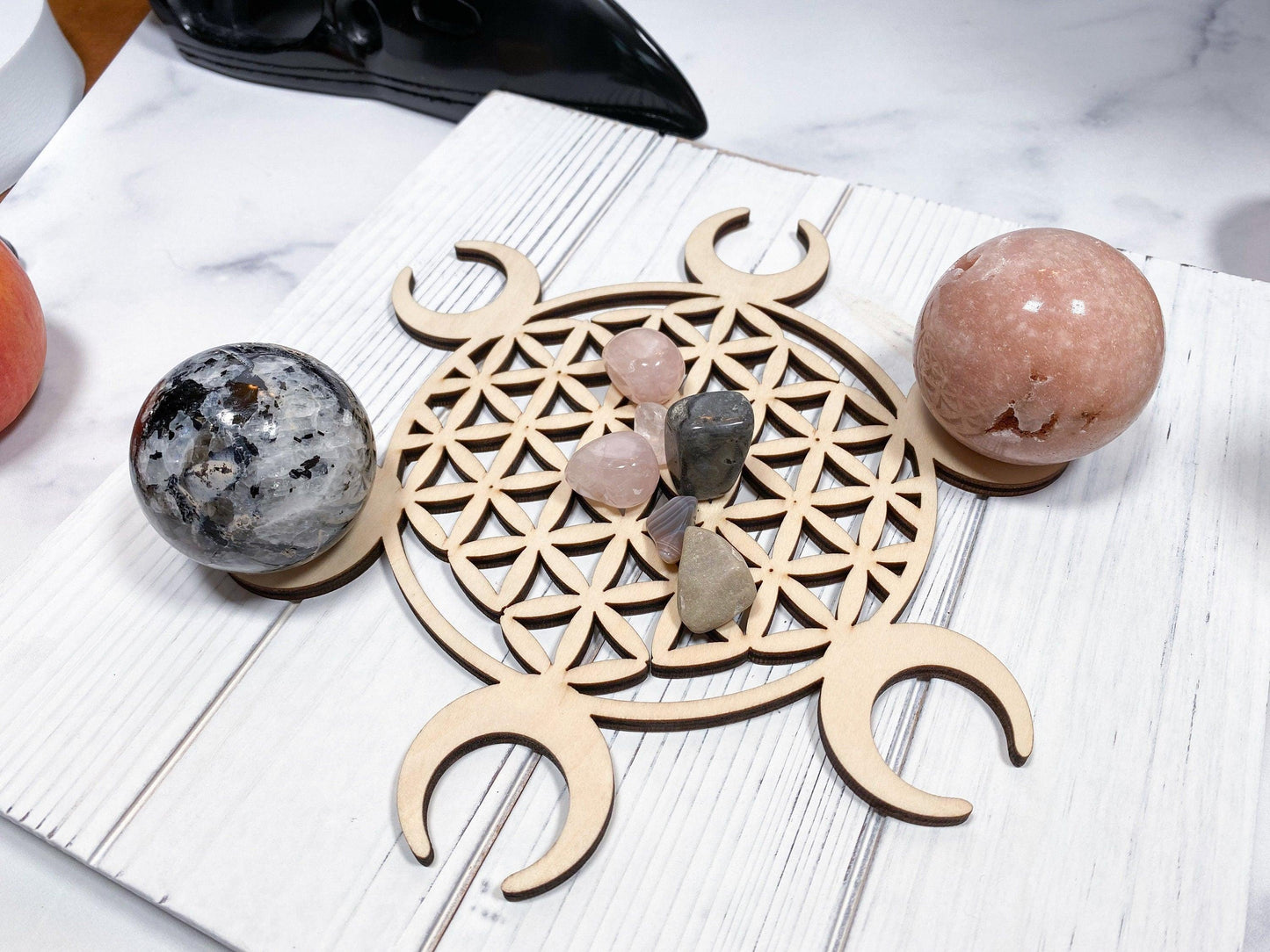 The Sacred Assorted Crystal Grids in Birch by The Crystalary, showcasing a stunning Flower of Life pattern with crescent moons, holds various crystal combinations. Two crystal spheres—one gray with black inclusions and the other pink—are elegantly placed beside the crystal grid on a white marble surface, creating an enchanting display of sacred geometry.