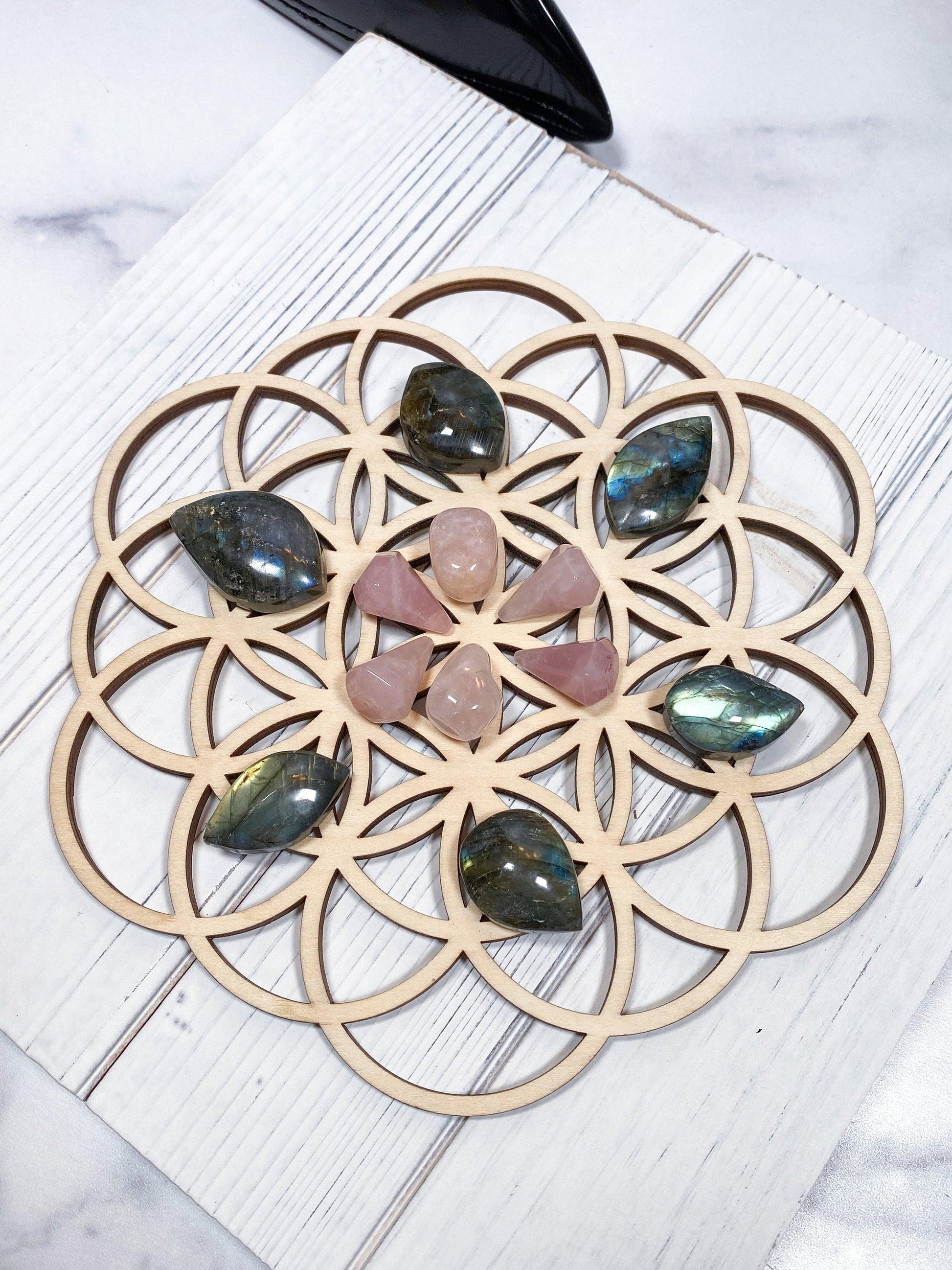 The Crystalary's Sacred Assorted Crystal Grids in Birch is beautifully arranged with polished stones in brown, pink, and green hues, meticulously placed to form a symmetrical design. This captivating grid featuring sacred geometry rests elegantly on a light birch surface.