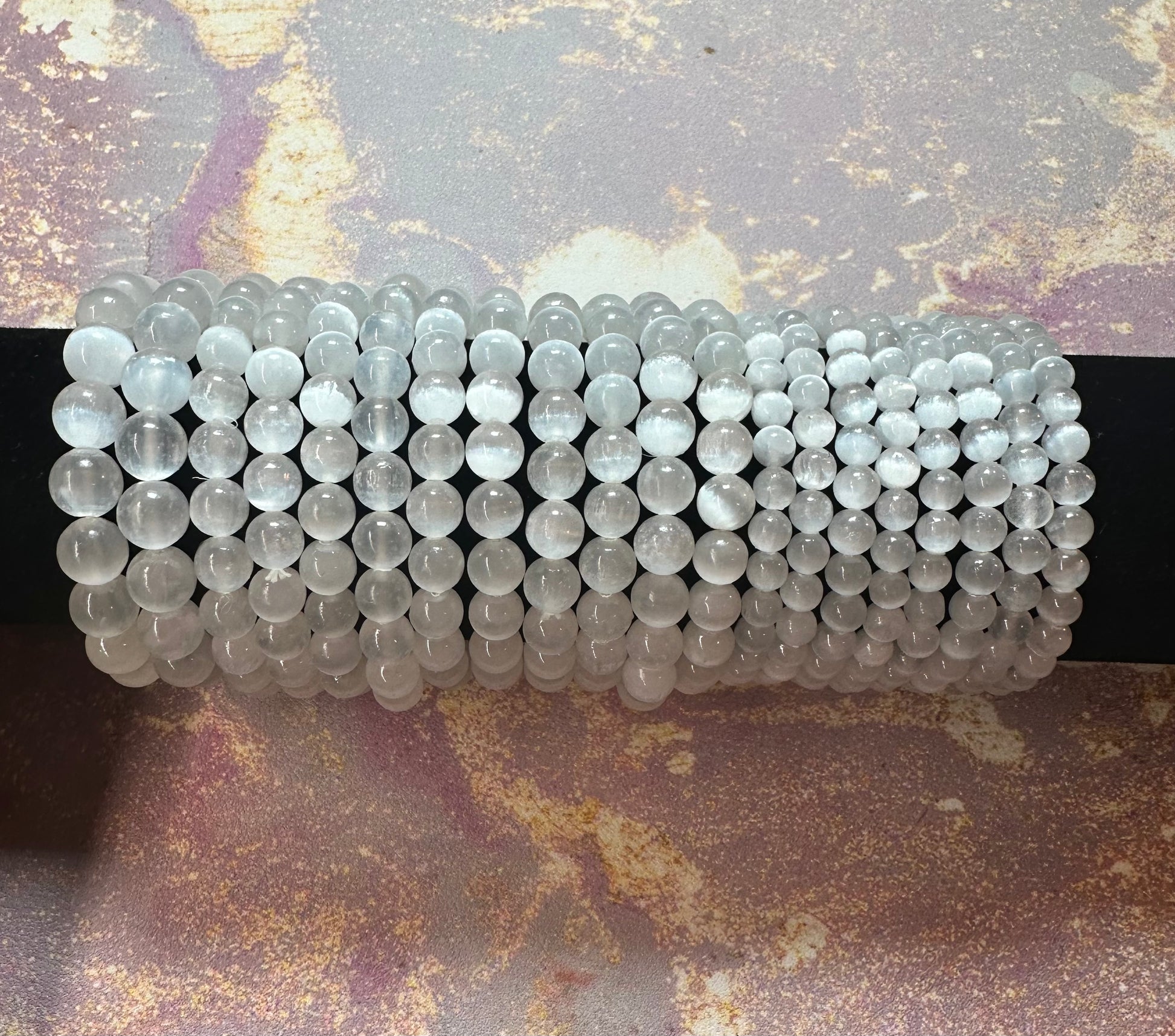 A row of ten identical Selenite Crystal Bracelets from The Crystalary is displayed on a black rod. The bracelets, made of round, translucent beads, are neatly stacked against a textured, marbled background in shades of pink and gray. These ethically sourced crystals are renowned for promoting mental clarity.