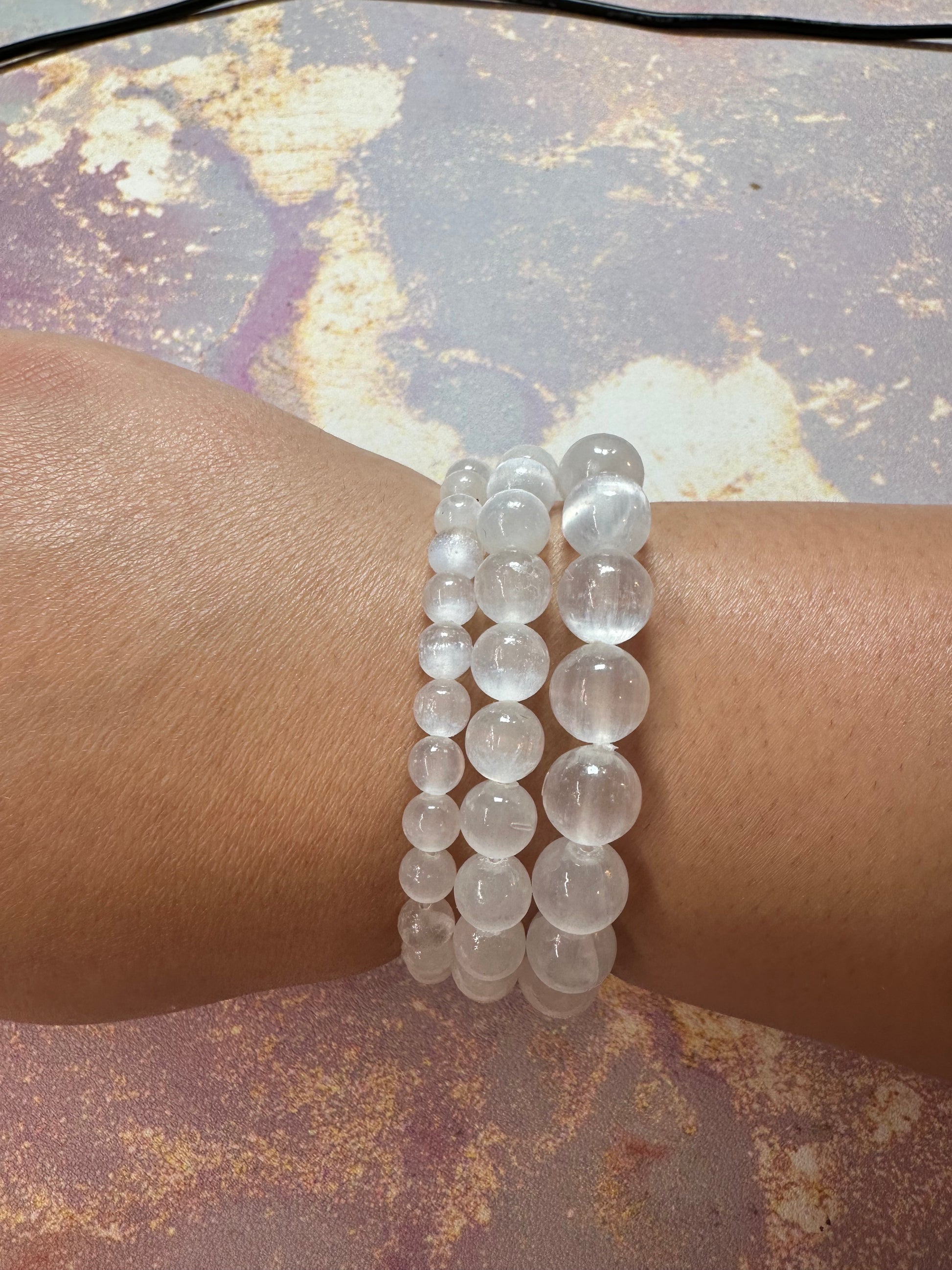 A wrist adorned with two Selenite Crystal Bracelets from The Crystalary, showcasing clear beads of varying sizes. The smaller bracelet features uniformly small beads, while the larger one displays medium-sized beads. The wrist's light skin tone contrasts against a textured pastel background that promotes peace through ethical crystal use.