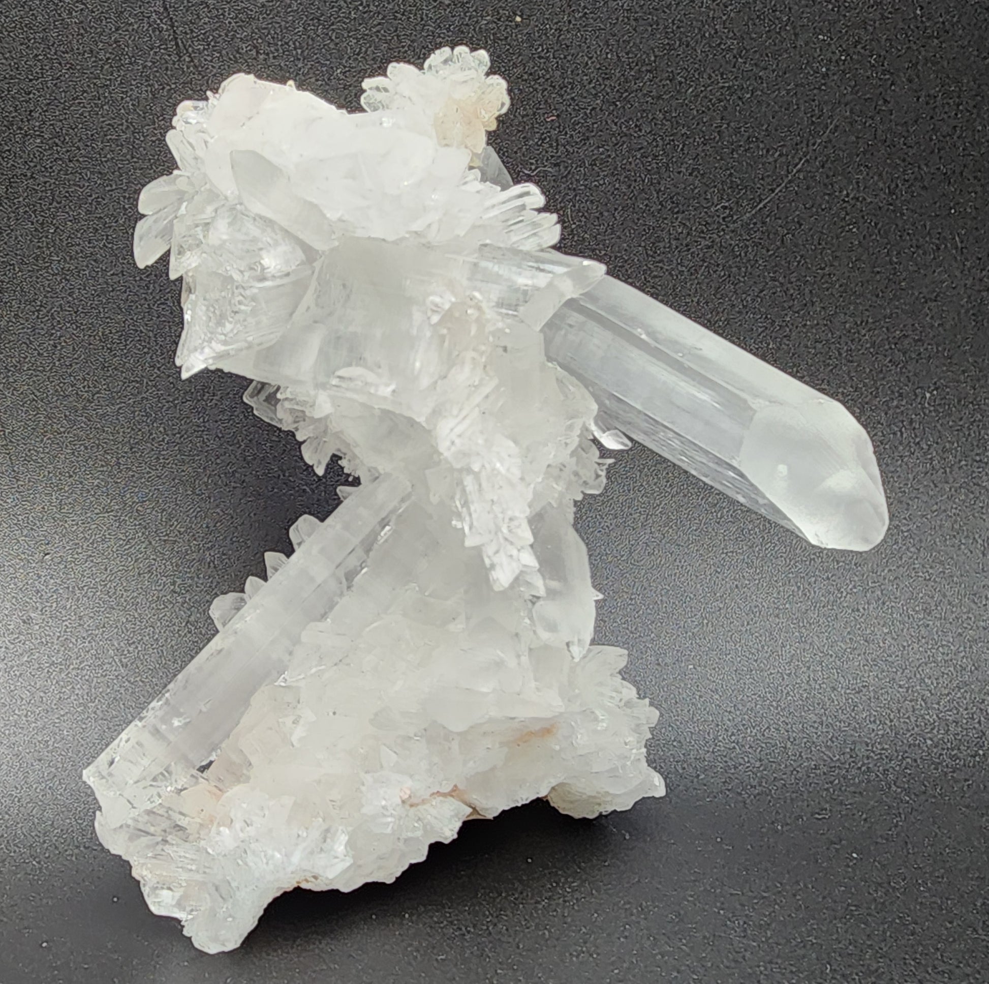 A cluster of clear, white Selenite crystals from the Naica Mine, Naica, Saucillo Municipality, Chihuahua, Mexico by The Crystalary on a black background. The formation features long, prismatic crystals pointing in various directions and interspersed with smaller, intricate crystal structures. The texture appears smooth and translucent.