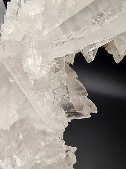 Close-up image of intersecting, translucent Selenite crystalline structures with sharp edges and layers, set against a dark background. The crystals exhibit a variety of angles, creating an intricate and detailed pattern reminiscent of formations found in the Naica Mine. This exquisite piece is part of The Crystalary's collection: Selenite- Naica Mine, Naica, Saucillo Municipality, Chihuahua, Mexico.