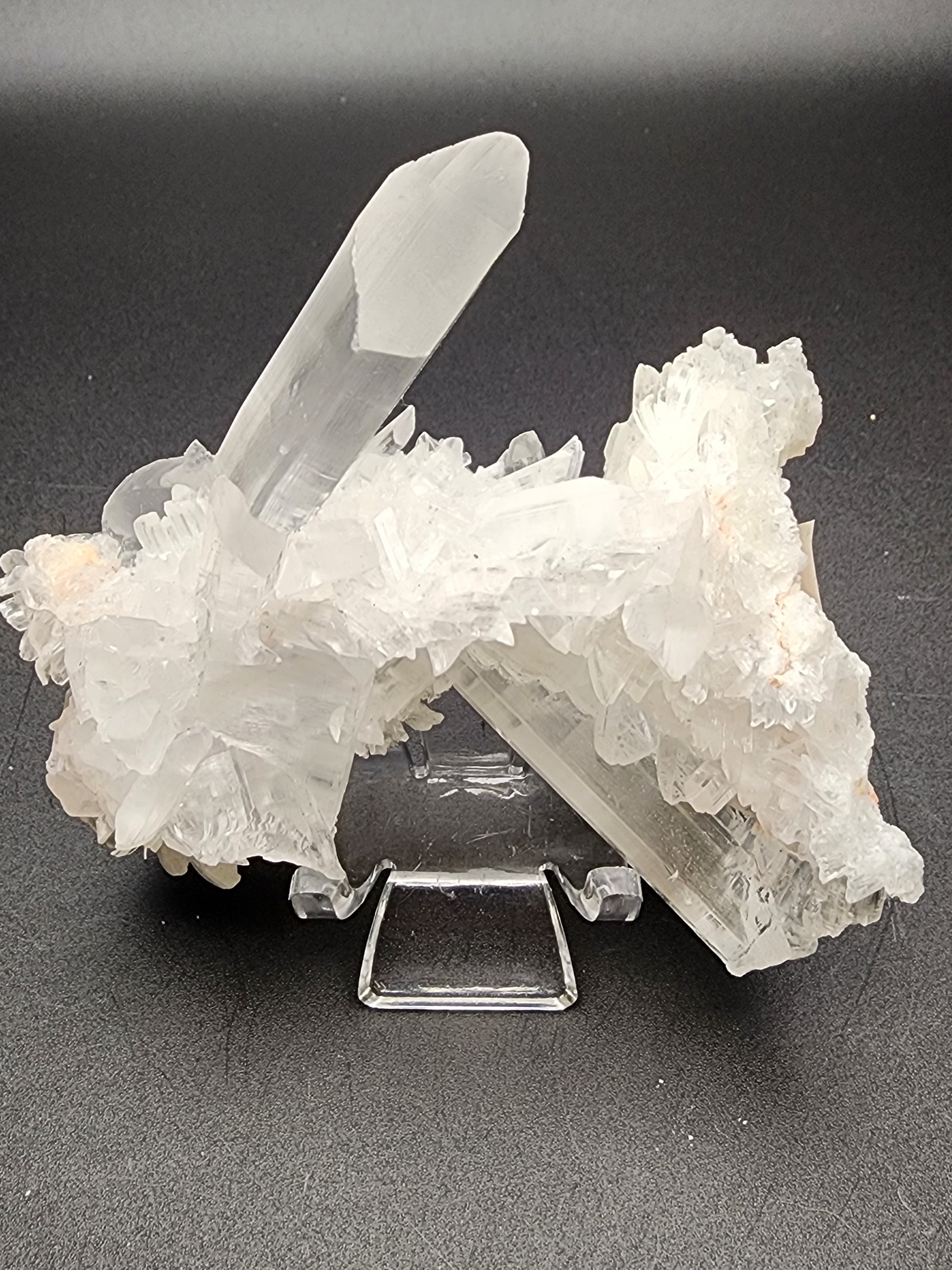 A cluster of Selenite crystals from the Naica Mine in Chihuahua, Mexico, displayed against a black background. These crystals from The Crystalary are mostly transparent with some white, opaque areas, and they form elongated, jagged shapes reminiscent of the formations found in Naica Mine. The cluster is elegantly displayed on a clear stand.