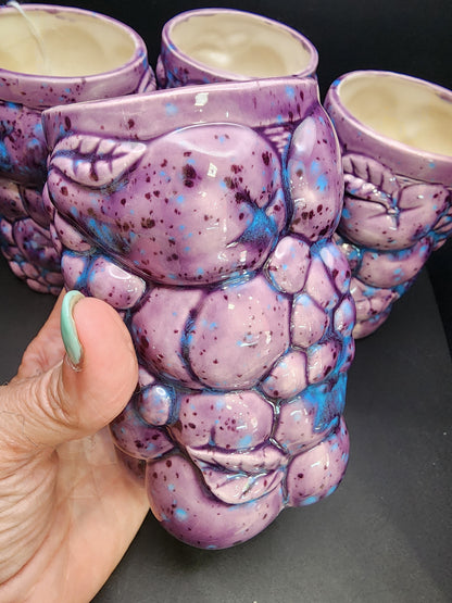 A hand holds a tumbler from The Crystalary's Set of 4 Inarco STYLE Ceramic Fruit Tumblers. The purple tumbler features a textured design resembling grapes, and there are three more identical tumblers in the background. Each glaze boasts speckles of blue and dark purple, giving the tumblers a vibrant and artistic appearance reminiscent of the iconic Inarco style.