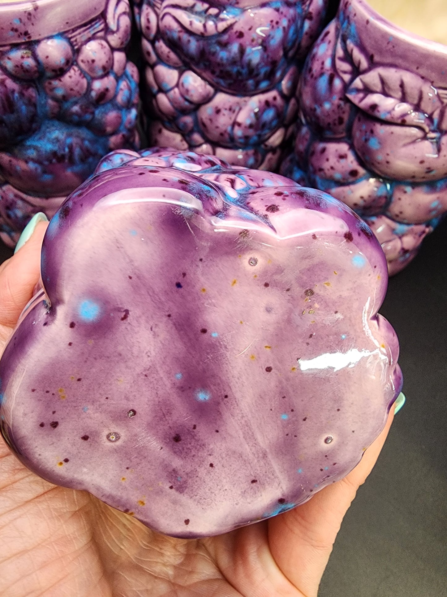 A hand holds a shiny, purple ceramic object with blue specks and subtle brown spots. In the background, three tumblers from The Crystalary's Set of 4 Inarco STYLE Ceramic Fruit Tumblers are visible, each featuring grape cluster designs with leaves and textures in beautiful purple hues with blue speckles. The nails, painted a turquoise color, add to the vibrant display.