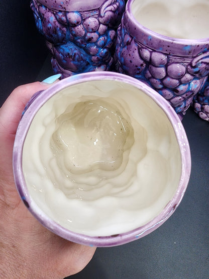 A hand holding a ceramic tumbler from The Crystalary's set of 4 Inarco STYLE Ceramic Fruit Tumblers, featuring a primarily purple exterior with blue speckles. The inside showcases a smooth, white, glazed finish with an organic, rippled effect at the bottom.