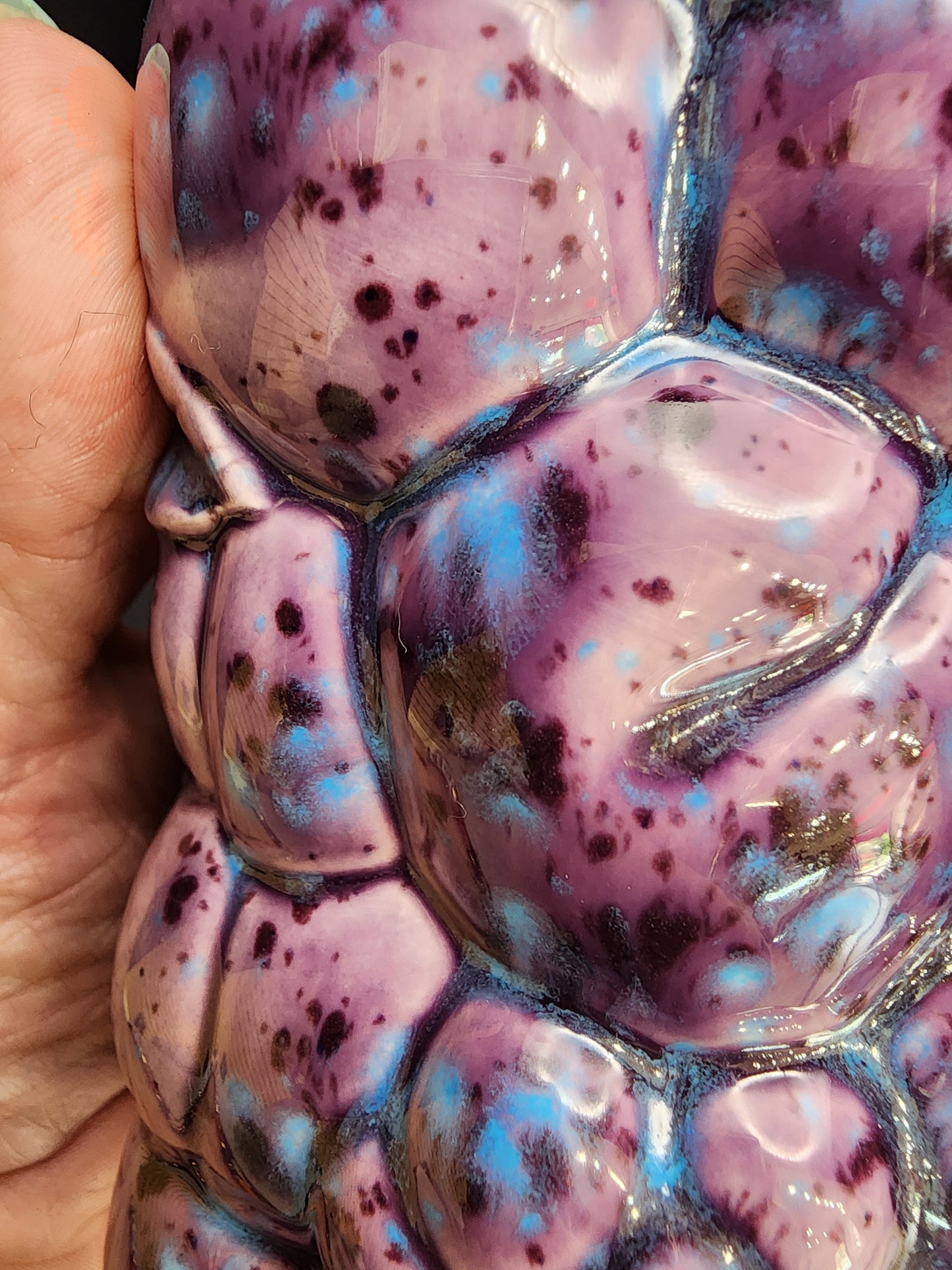 A close-up of a hand-painted ceramic object with a glossy, bumpy texture reminiscent of the Inarco style by The Crystalary. Primarily purple with blue and black speckles, it resembles one of the purple fruit ceramic tumblers from their Set of 4. A hand, slightly out of focus, holds part of the polished, reflective surface.