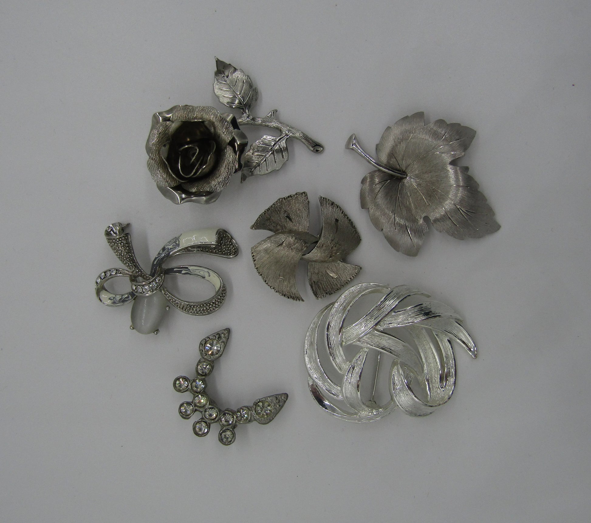 A set of five vintage silver tone pins from The Crystalary, including designs such as a rose, a leaf, a bow, an abstract swirl with rhinestones, and a intricately patterned butterfly-like shape. Perfect for mix and match to elevate any outfit with these eclectic brooches.