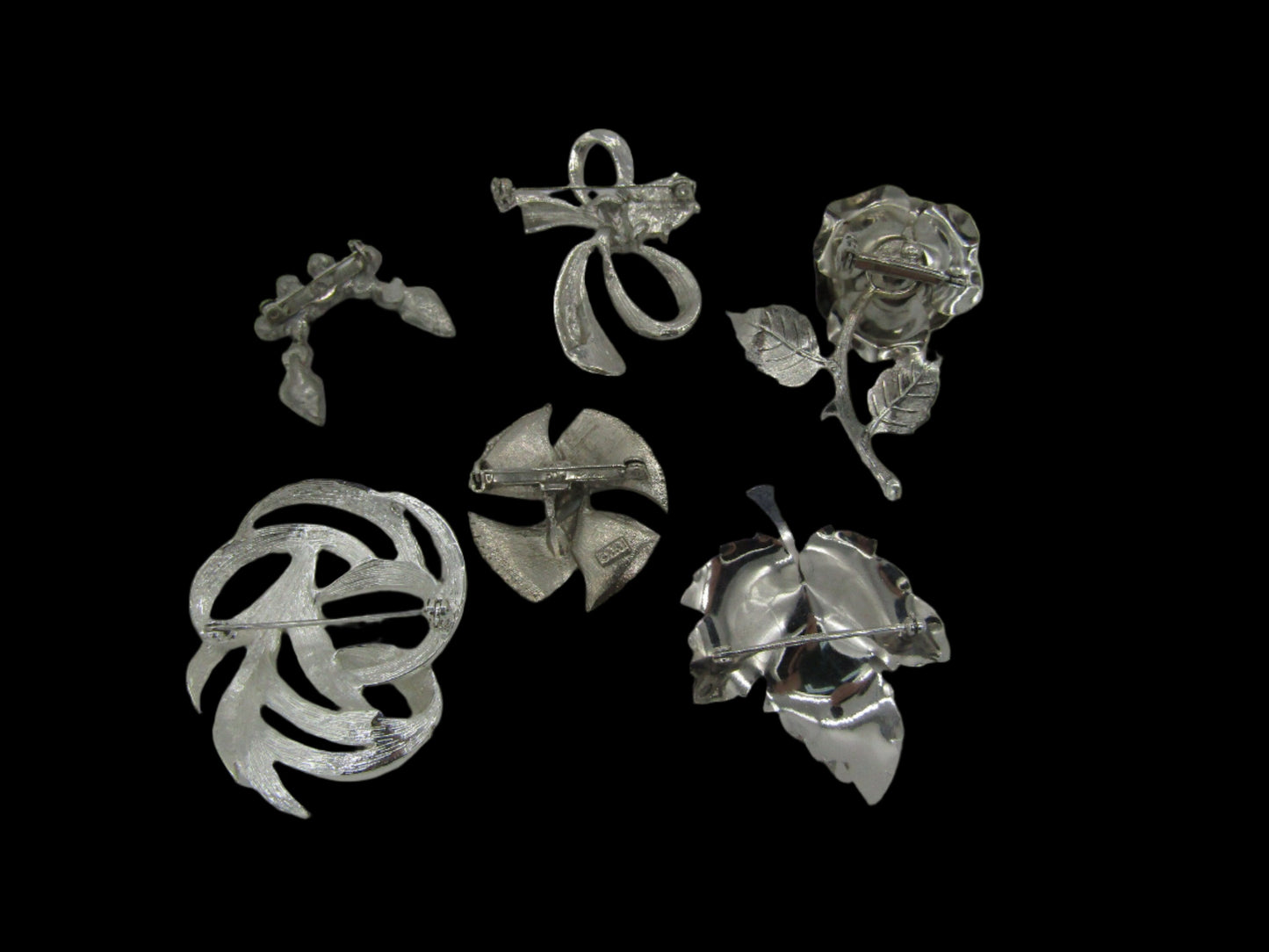 A black background displays five vintage brooches from The Crystalary's Set of 5 Vintage Silver Tone Pins/Brooches collection. The designs include a ribbon shape, a leaf with fruit, an abstract pattern, a tulip, and a rose with leaves. Each silver-tone brooch showcases intricate details and craftsmanship reminiscent of modern 70s aesthetics, enhanced with rhinestones.