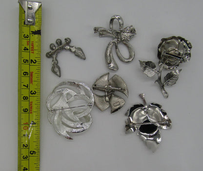 The Crystalary's Set of 5 Vintage Silver Tone Pins/Brooches, featuring rhinestones and designed in abstract modern shapes from the 70s—including leaves, a flower, a bow, and a fan—are displayed alongside a yellow measuring tape that indicates their sizes range from approximately 1 to 3 inches.