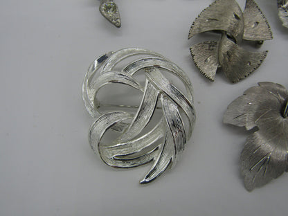 A close-up image showcases a Set of 5 Vintage Silver Tone Pins/Brooches from The Crystalary, arranged against a white background. The central brooch boasts an intricate, layered design with overlapping curved elements, while the surrounding pins offer a delightful mix of abstract patterns and textures.