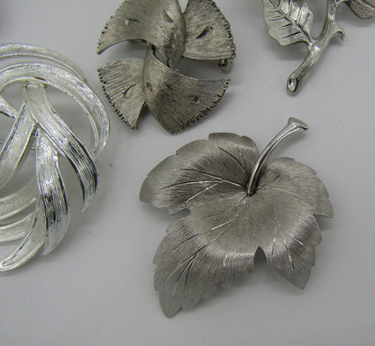 A set of 5 vintage silver tone pins and brooches from The Crystalary, artistically arranged on a white background, showcasing modern abstract designs from the 70s. Featured designs include a complex knot, multiple ginkgo leaves, and a large intricately detailed maple leaf. Each piece boasts textured surfaces adorned with rhinestones that highlight their meticulous craftsmanship.