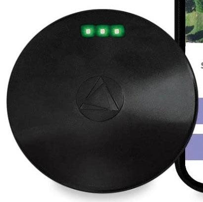 A round black electronic device with three small green LED lights on the top and a subtle triangular logo in the center, resembling The Crystalary's Shipping Add On: Theft/Loss Recovery GPS Tracker. The edge of a smartphone is partially visible on the right side of the image.