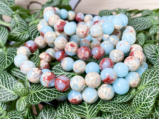 A close-up image of The Crystalary's Shoushan Jade Crystal Bracelets, showcasing round beads in shades of light blue, white, and red. The bracelets are elegantly displayed on a bed of green leafy plants with white veining, offering a striking contrast to the natural background.