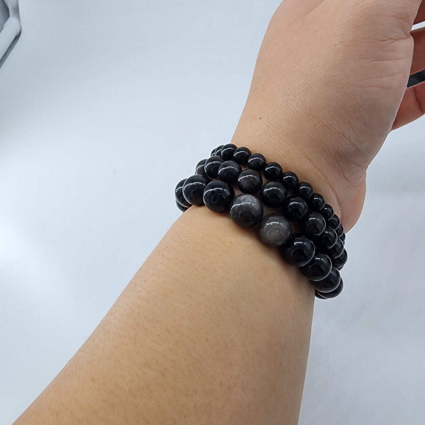 A close-up image of a person's wrist adorned with three Silver Sheen Obsidian Crystal Bracelets from The Crystalary, crafted with 7-inch elastic bands, stacked elegantly against a plain white background. The shiny, polished beads vary slightly in size. The person holds their wrist at a slight upward angle, exuding an air of sharpening intellect.