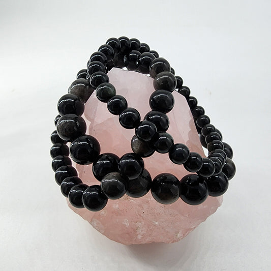 The Silver Sheen Obsidian Crystal Bracelet from The Crystalary is elegantly draped over a rose quartz crystal, showcasing a stunning contrast between the pale pink stone and the glossy dark beads. This visually striking composition is set against a simple, clean white background, highlighting the beauty of both elements.