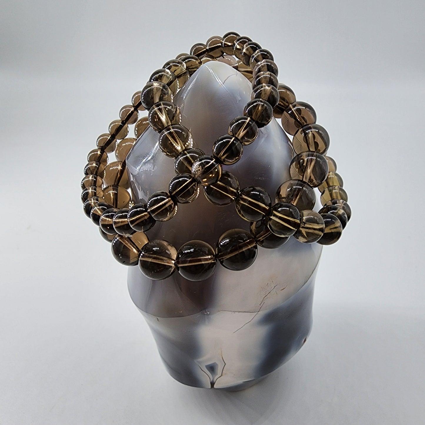 A polished Smoky Quartz Crystal from The Crystalary stands upright, exuding positive energy. It is adorned with multiple strands of round, brownish beads intricately wrapped around it in a lattice-like pattern. The background is plain white.