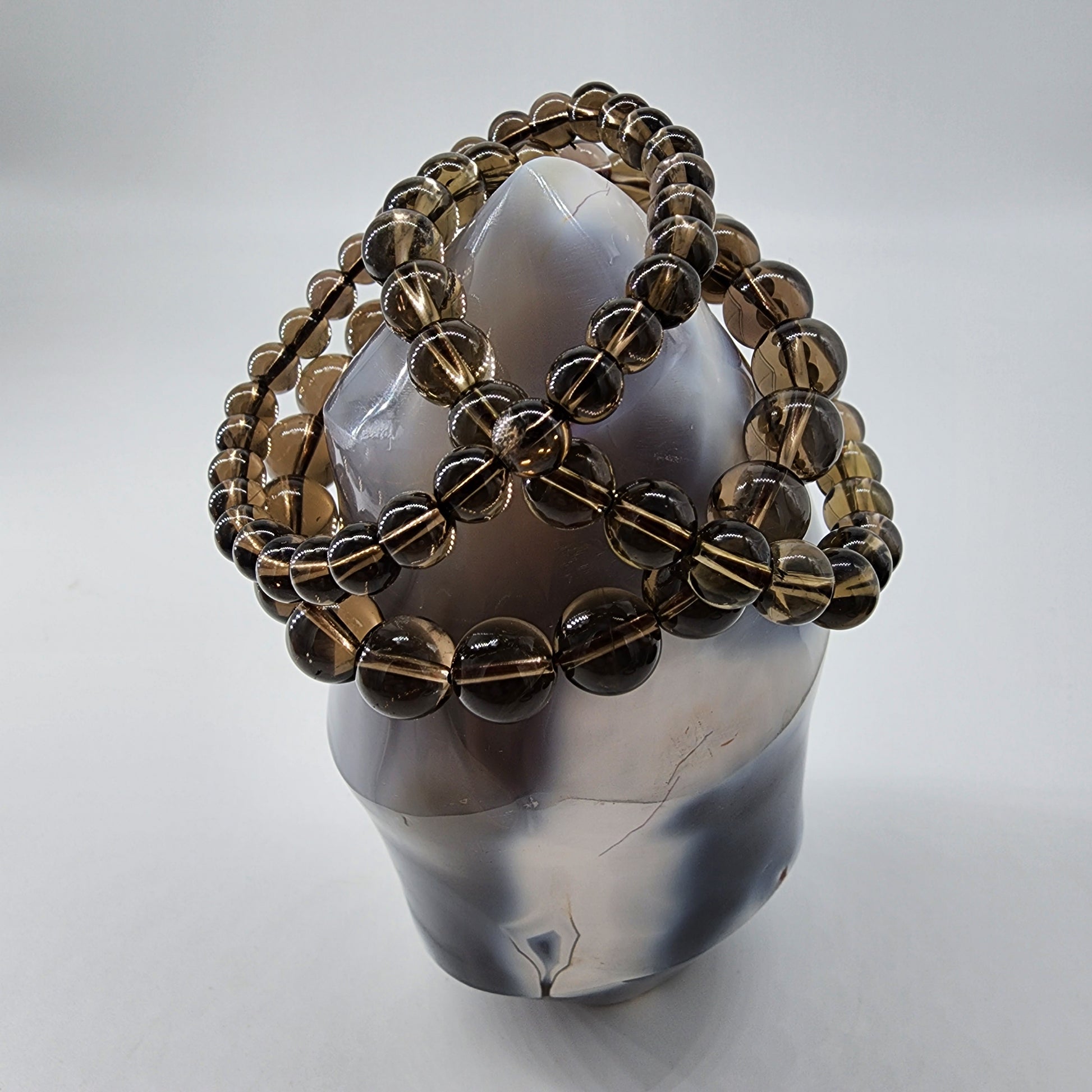 A polished Smoky Quartz Crystal from The Crystalary stands upright, exuding positive energy. It is adorned with multiple strands of round, brownish beads intricately wrapped around it in a lattice-like pattern. The background is plain white.