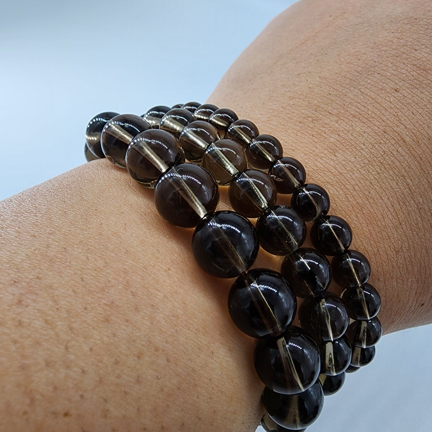 A close-up image of a wrist adorned with the Smoky Quartz Crystal Bracelet by The Crystalary showcases multiple strands of round, smoky quartz beads in a smoky grey color. The glossy, smooth beads are arranged tightly around the wrist in a layered fashion, promising to disperse fear and invite positive energy. The background is plain and light-colored.