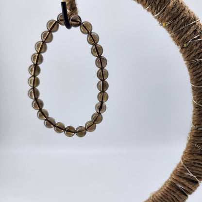 A close-up photo of The Crystalary's Smoky Quartz Crystal Bracelet, featuring round brown translucent beads, hanging on a circular jute-wrapped structure. The slightly blurred grayish-white background enhances the intricate details of the bracelet, which is believed to disperse fear and invite positive energy.