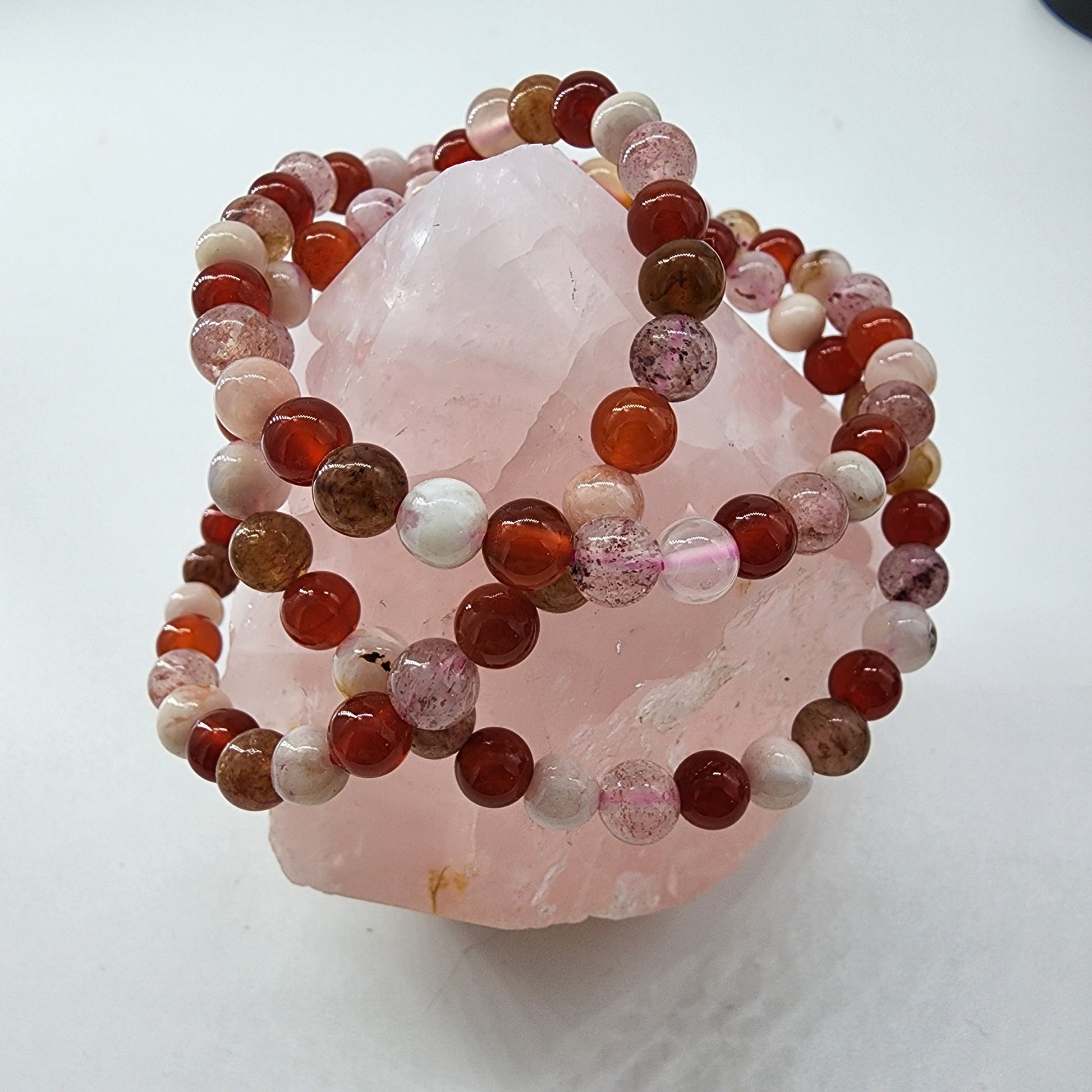 A pink rose quartz crystal with several Strawberry Quartz, Carnelian, and Flower Agate Crystal Bracelets draped over it exudes emotional warmth. The bracelets from The Crystalary feature round beads in various shades of pink, red, and brown, with some beads being translucent or speckled. The creative setup is against a plain off-white background.