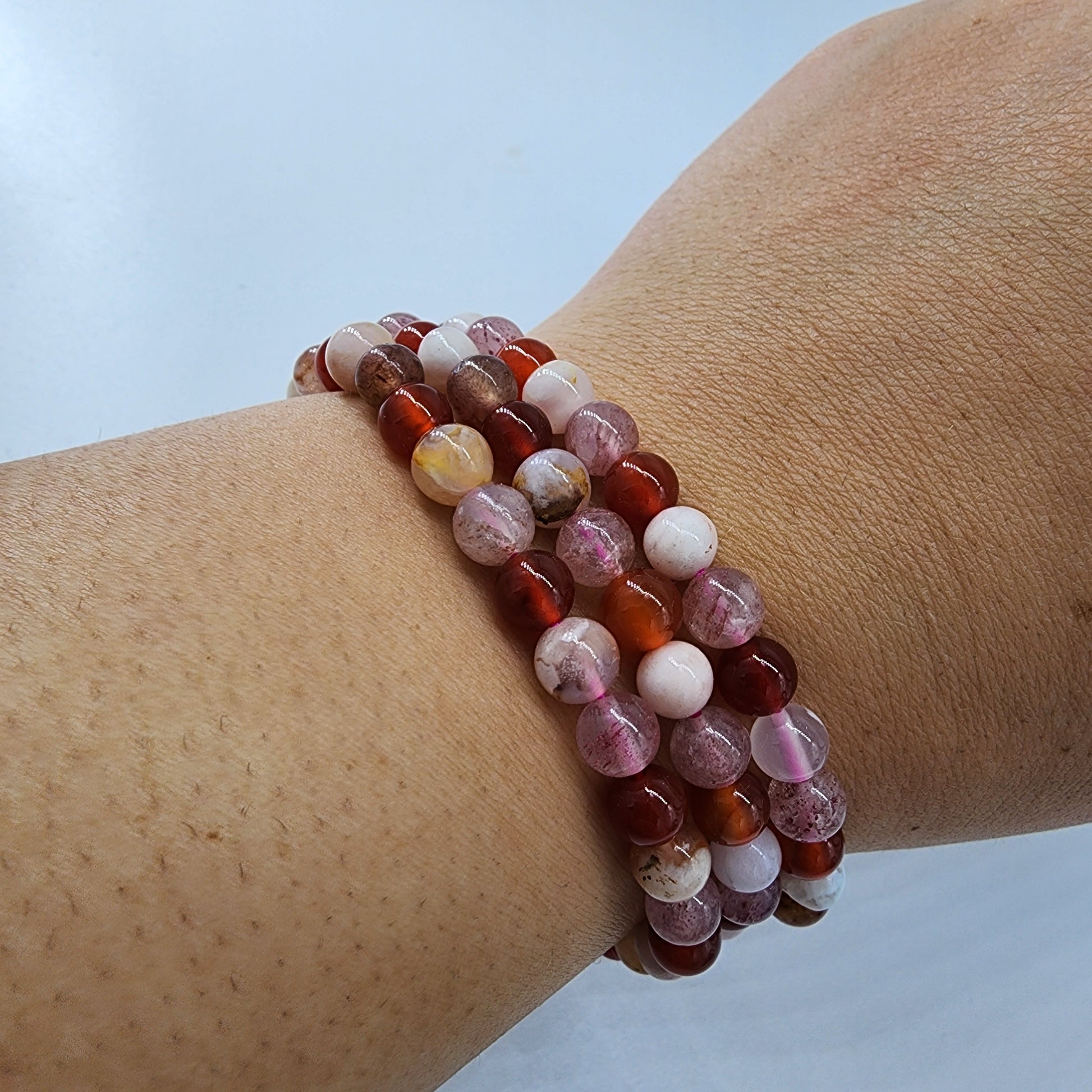 A close-up of a wrist adorned with The Crystalary's Strawberry Quartz, Carnelian, and Flower Agate Crystal Bracelets, radiating emotional warmth. The beads vary in shades of orange, red, white, and pink with unique mottled patterns. The background is plain, highlighting the light skin tone of the wrist.