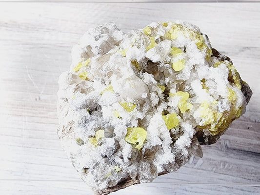 A close-up image of Sulfur from Maybee, Michigan by The Crystalary, showcasing a mix of white, yellow, and grey crystal formations. The 107 g mineral rock rests on a light-colored, textured surface.