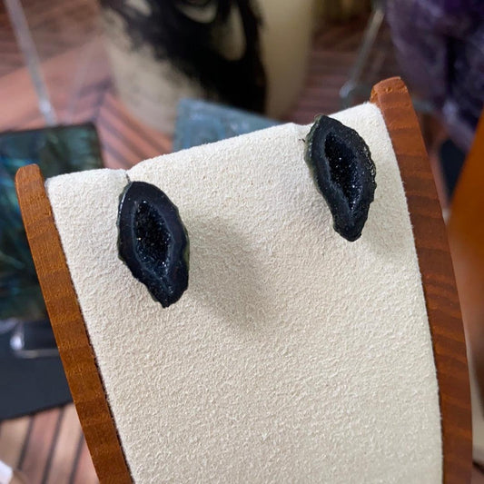 A close-up of a pair of Tobasco Geode Stud Earrings by The Crystalary, displayed on a light-colored, textured stand with wooden edges. The unique small geodes are unevenly shaped with a glittery interior, highlighting their natural, raw appearance. The background is blurred.