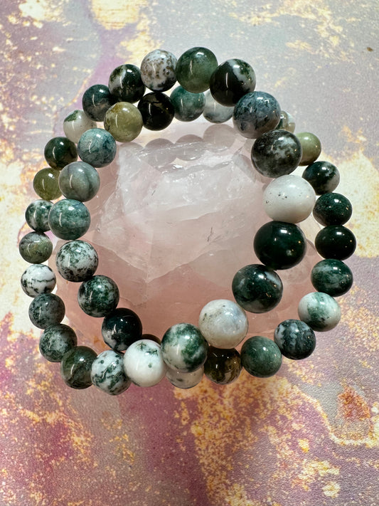 The Tree Agate Crystal Bracelet by The Crystalary, made of green and white marbled stones, is displayed on a large, translucent pink crystal. The backdrop features a textured surface in pastel shades of pink, yellow, and purple, creating an inviting scene for those seeking ethical crystal decor or elegant gemstone bracelets.