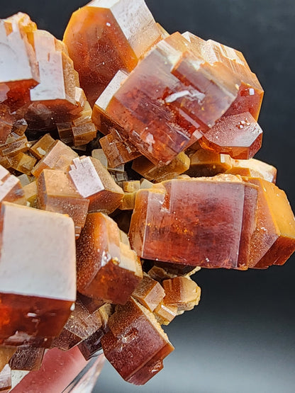 A close-up showcases a cluster of translucent, reddish-brown Vanadinite crystals from The Crystalary, originating from the Mibladen mining district in Midelt Province, Drâa-Tafilalet Region, Morocco (sku 3076). These tightly packed cubic crystals boast smooth surfaces and reflective edges, forming an intricate geometric pattern against a dark background.