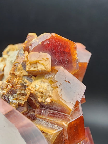 Close-up image of a cluster of Vanadinite crystals from The Crystalary, featuring a glossy surface and originating from the Mibladen mining district in Midelt Province, Drâa-Tafilalet Region, Morocco (SKU 3076). Set against a dark background, these reddish-brown crystals exhibit sharp edges and varying shades of amber and brown, showcasing their intricate structure.