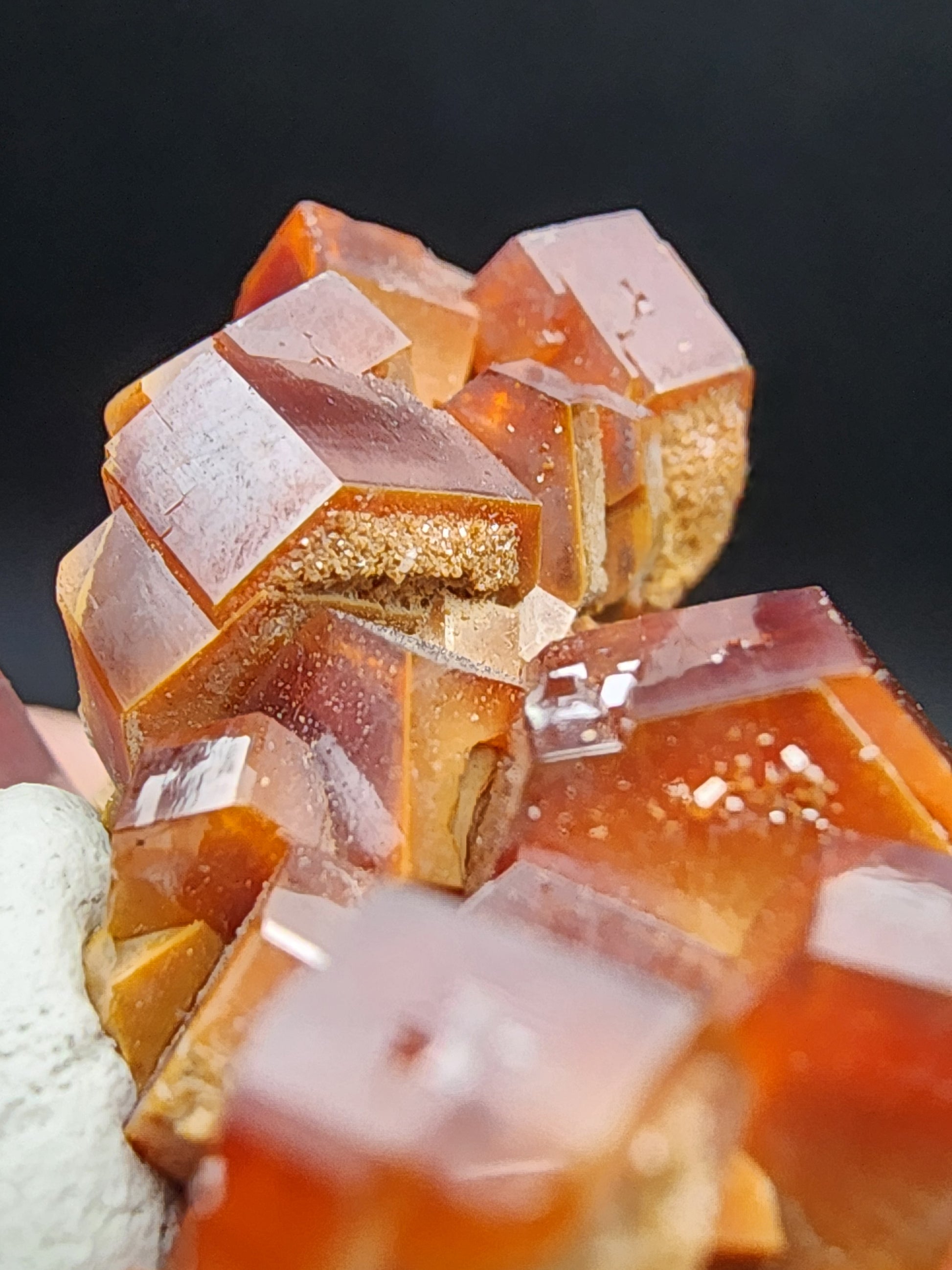 This is a close-up of vanadinite crystals from the prestigious Mibladen mining district in Morocco. These reddish-brown cubic crystals, catalogued as SKU 3076 by The Crystalary, are set against a contrasting beige rock and exhibit shiny surfaces that beautifully stand out against a dark background. Their geometric shapes form an eye-catching cluster.