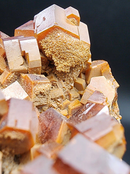 A close-up image showcases a cluster of brown, cubic vanadinite crystals from The Crystalary (sku 3076), sourced from the Mibladen mining district in Midelt Province, Drâa-Tafilalet Region, Morocco. The crystals feature a rough, granular texture on some surfaces and are set against a dark background. Their varying sizes and orientations form an intricate geometric pattern.