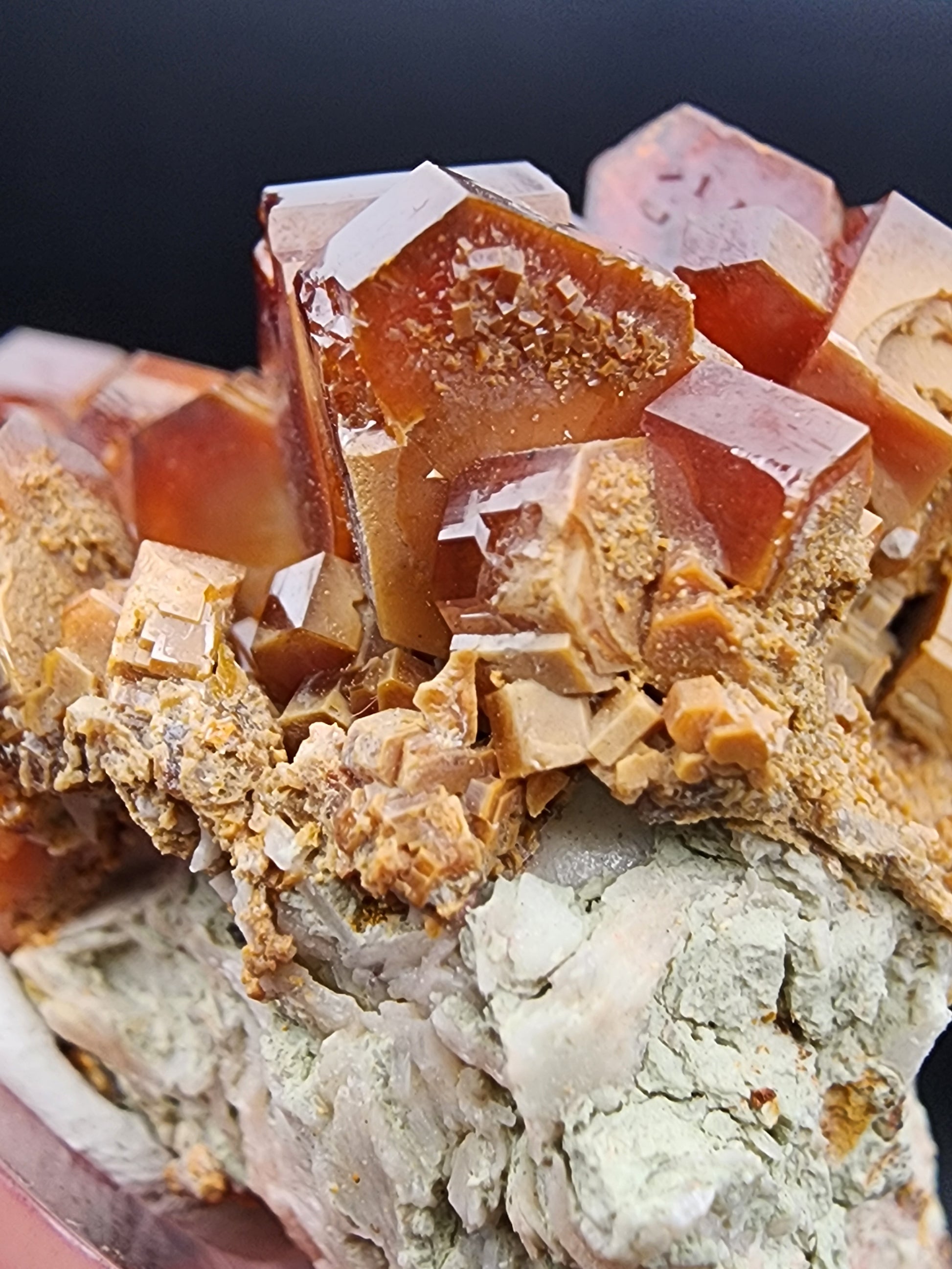 Close-up view of The Crystalary's Vanadinite crystals (SKU 3076) from the renowned Mibladen mining district in the Midelt Province, Drâa-Tafilalet Region, Morocco. These reddish-brown crystals feature smooth surfaces and sharp edges, beautifully embedded in a rough, light-colored mineral matrix. The vivid hues of the striking cubic formations contrast elegantly with the irregular texture of the surrounding rock.