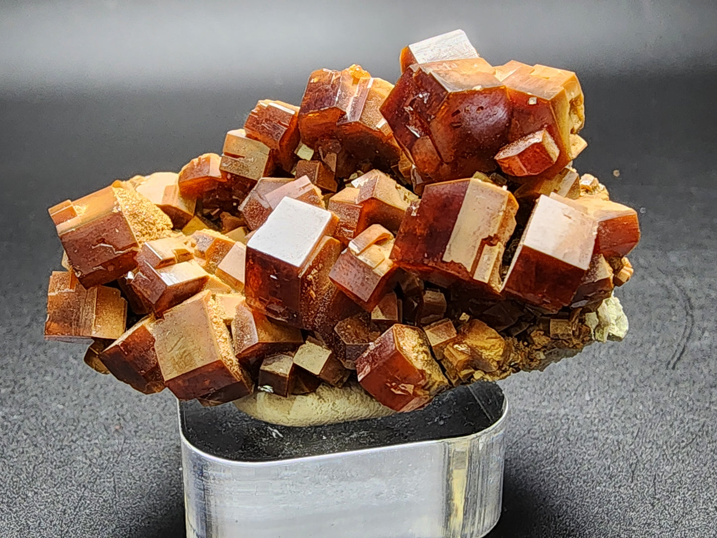 The Crystalary presents a captivating cluster of reddish-brown vanadinite crystals, product SKU 3076, sourced from the renowned Mibladen mining district in Midelt Province, Drâa-Tafilalet Region, Morocco. These cube-like crystals feature smooth surfaces and sharp edges and are arranged artistically in various orientations against a dark background on a small stand.