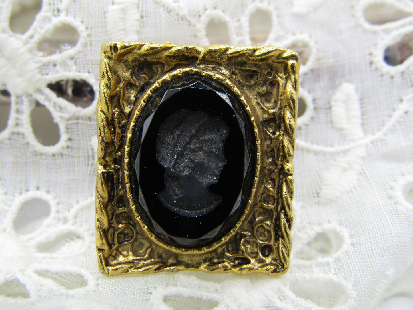 A Vintage Black Glass Reverse Relief Cameo Style Brooch/Pin Grecian lady by The Crystalary features a black oval center with the silhouette of a woman's profile. Its ornate gold frame, adorned with intricate designs and patina, is set against a delicate lace background, exuding the elegance of a Grecian style pin.