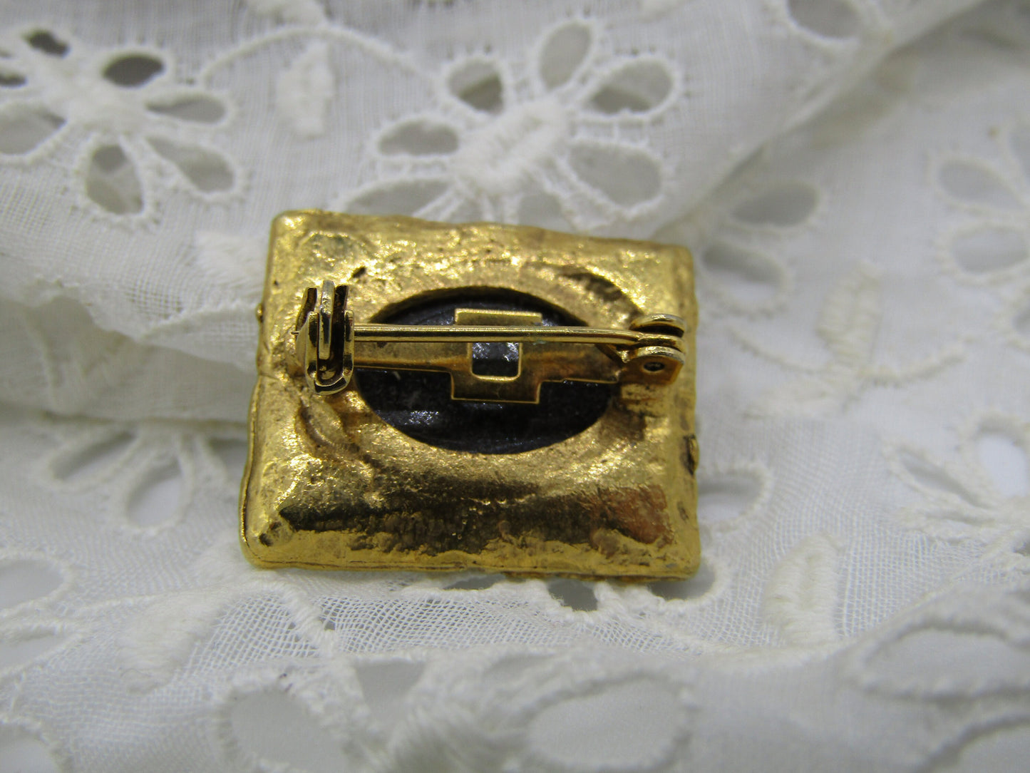 A small, square gold Vintage Black Glass Reverse Relief Cameo Style Brooch/Pin Grecian lady from The Crystalary, featuring a black circular center with an intricately detailed Grecian design, is placed on an intricately patterned white lace fabric. The brooch includes a simple mechanism on the back for fastening.