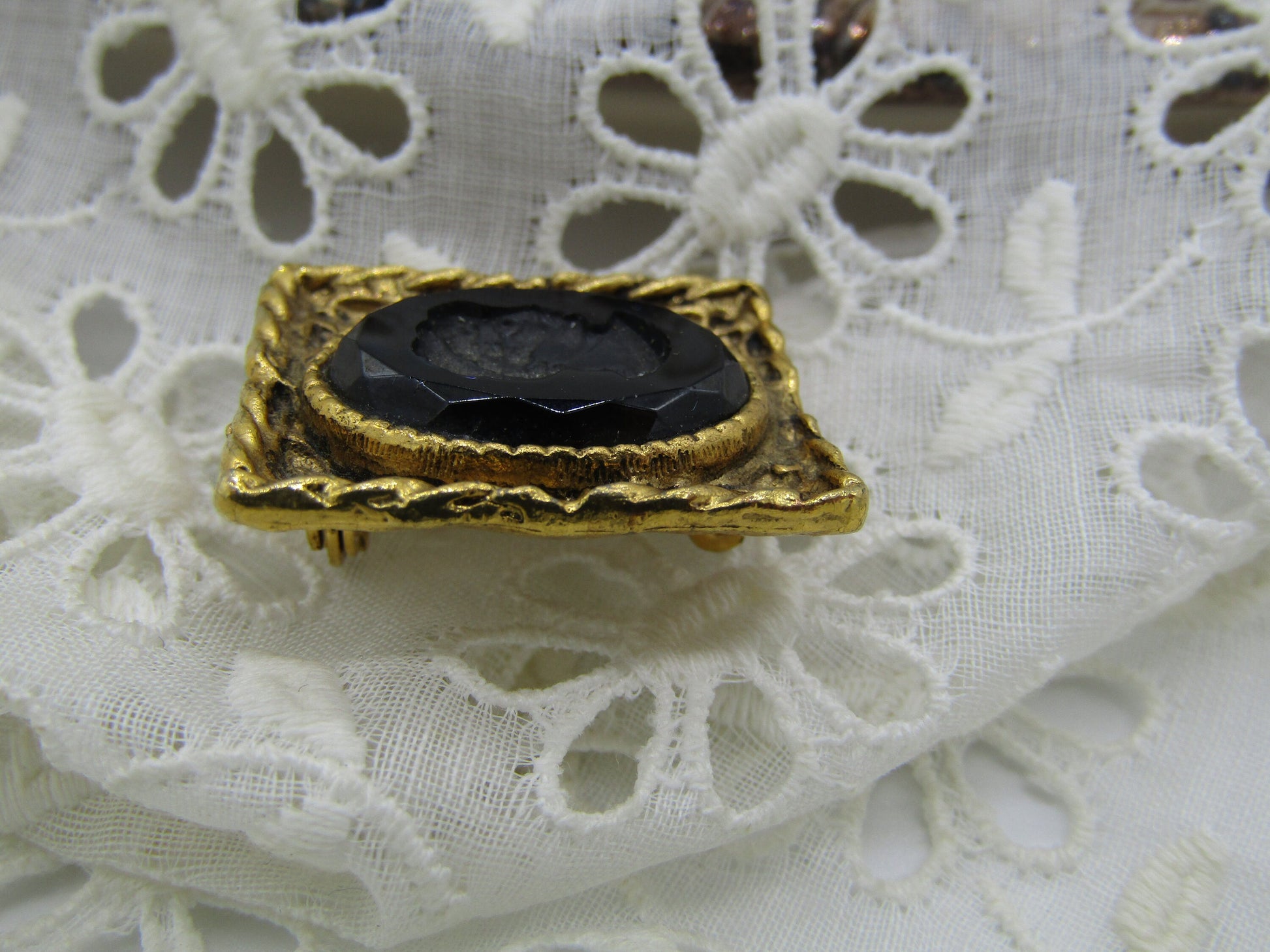 A gold brooch from The Crystalary, featuring a detailed Grecian lady design and a large black gemstone in the center, is set against a piece of white lace fabric with a floral pattern. The Vintage Black Glass Reverse Relief Cameo Style Brooch/Pin includes an intricately textured gold frame.