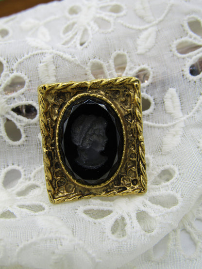 A Vintage Black Glass Reverse Relief Cameo Style Brooch/Pin featuring a Grecian lady from The Crystalary, with an intricate border surrounding a black oval center showcasing a profile of a person's head. This elegant pin is beautifully displayed on white lace fabric.