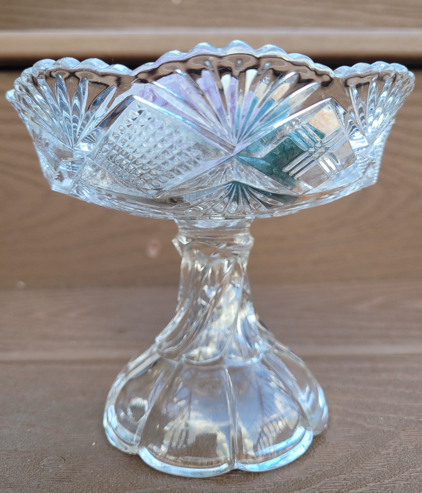 A stunning piece from The Crystalary, the Vintage EAPG Pedestal Pressed Glass Dish features intricate cut patterns and sits gracefully on a twisted pedestal base. The dish is adorned with a scalloped edge and a starburst design, beautifully showcasing its detailed craftsmanship against a wooden surface background.